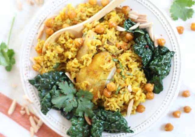 Plate with turmeric rice and chicken with a side of sauteed kale garnished with crispy chickpeas, toasted almonds and cilantro