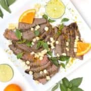 White plate with flank steak and corn, with limes, oranges and basil around the plate