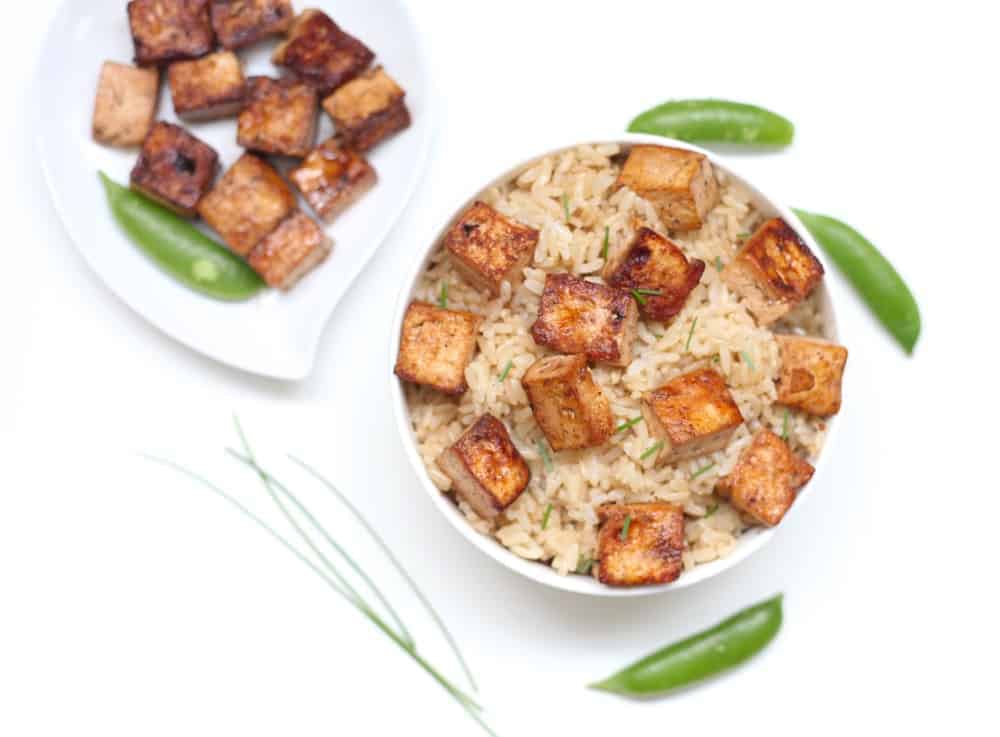 Seared tofu over brown rice in white bowl surrounded by snap peas and chives