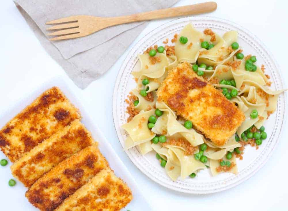 Slices of crispy, browned tofu fried in breadcrumbs on a white plate with buttered egg noodles and green peas