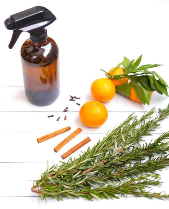 A glass spray bottle filled with natural kitchen cleaner on a white background surrounded by oranges, rosemary cloves and cinnamon sticks