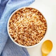 A bowl of cooked farro on a white background with a blue towel and small bamboo spoon