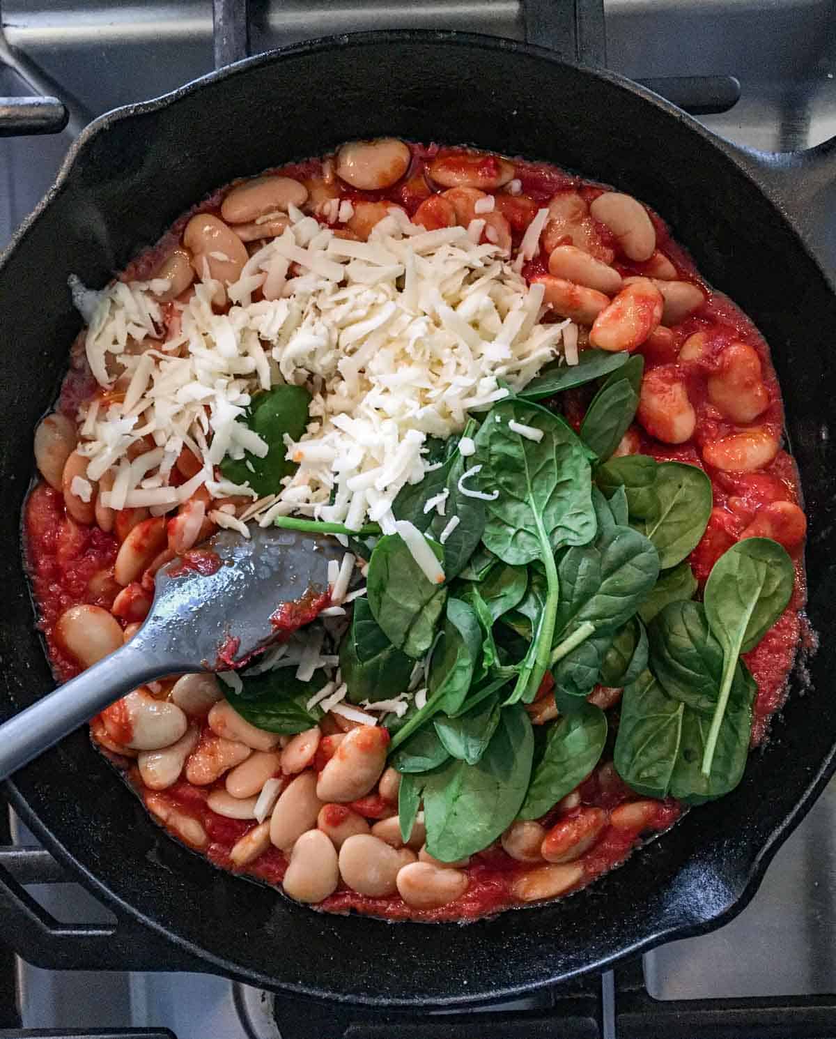 cast iron skillet filled with canned butter beans, tomato paste, spinach and grated cheese