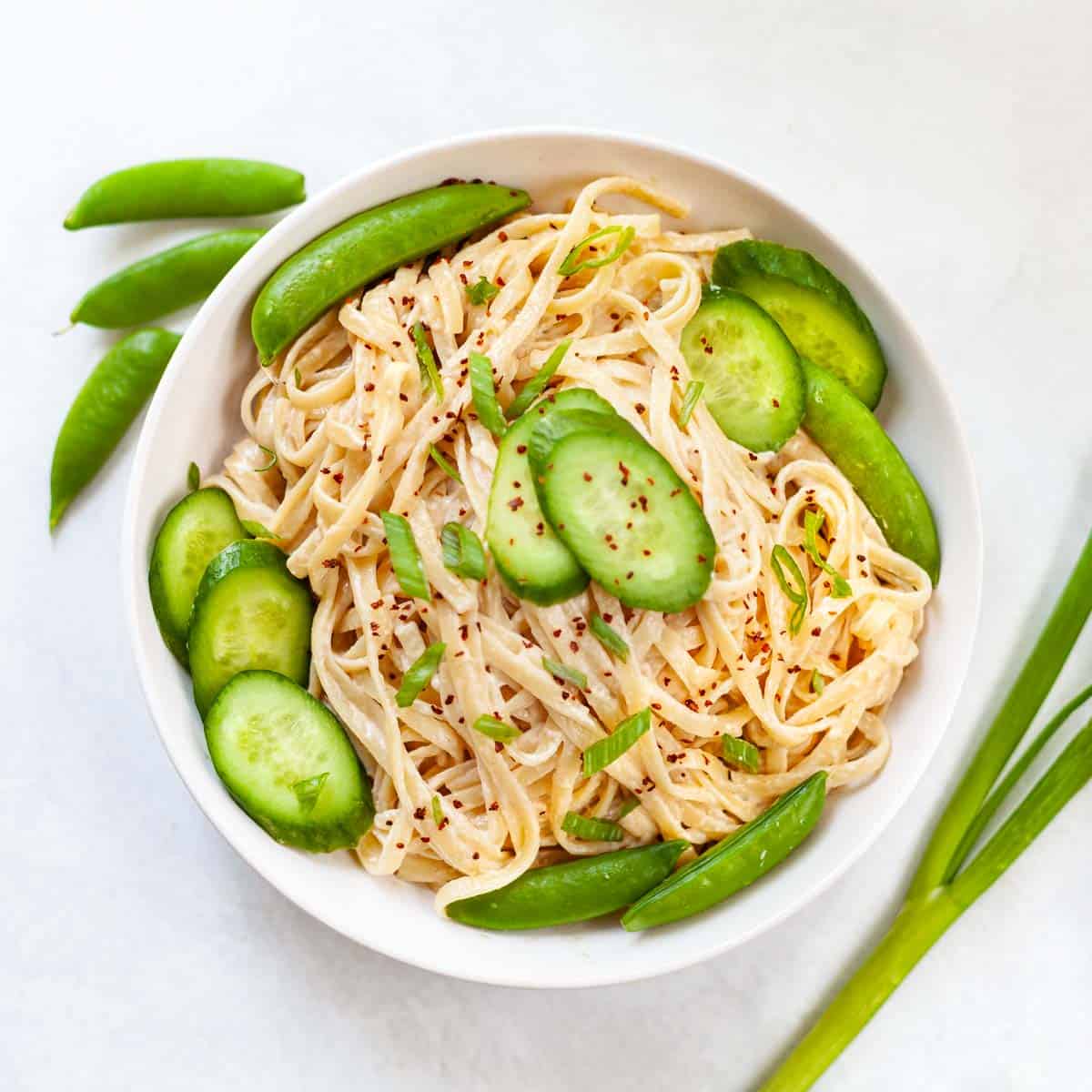 white bowl with fettucine tossed in a tahini dressing garnished with sliced cucumber and snap peas