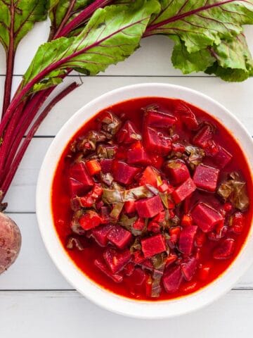 a bowl of beet soup next to a raw beet with greens attached