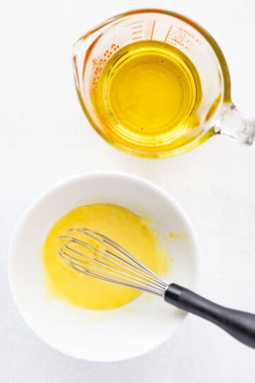 a bowl of aioli with a whisk next to a measuring cup of oil