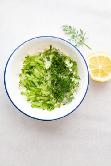 half of a lemon next to a bowl of sour cream, fresh herbs and grated cucumber