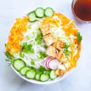 a bowl of cold rice noodles cucumber, cabbage, carrots, green onion and tofu next to a small bowl of soy sauce dressing