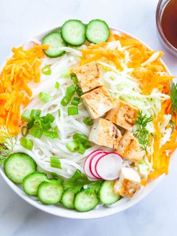 a bowl of cold rice noodles cucumber, cabbage, carrots, green onion and tofu next to a small bowl of soy sauce dressing