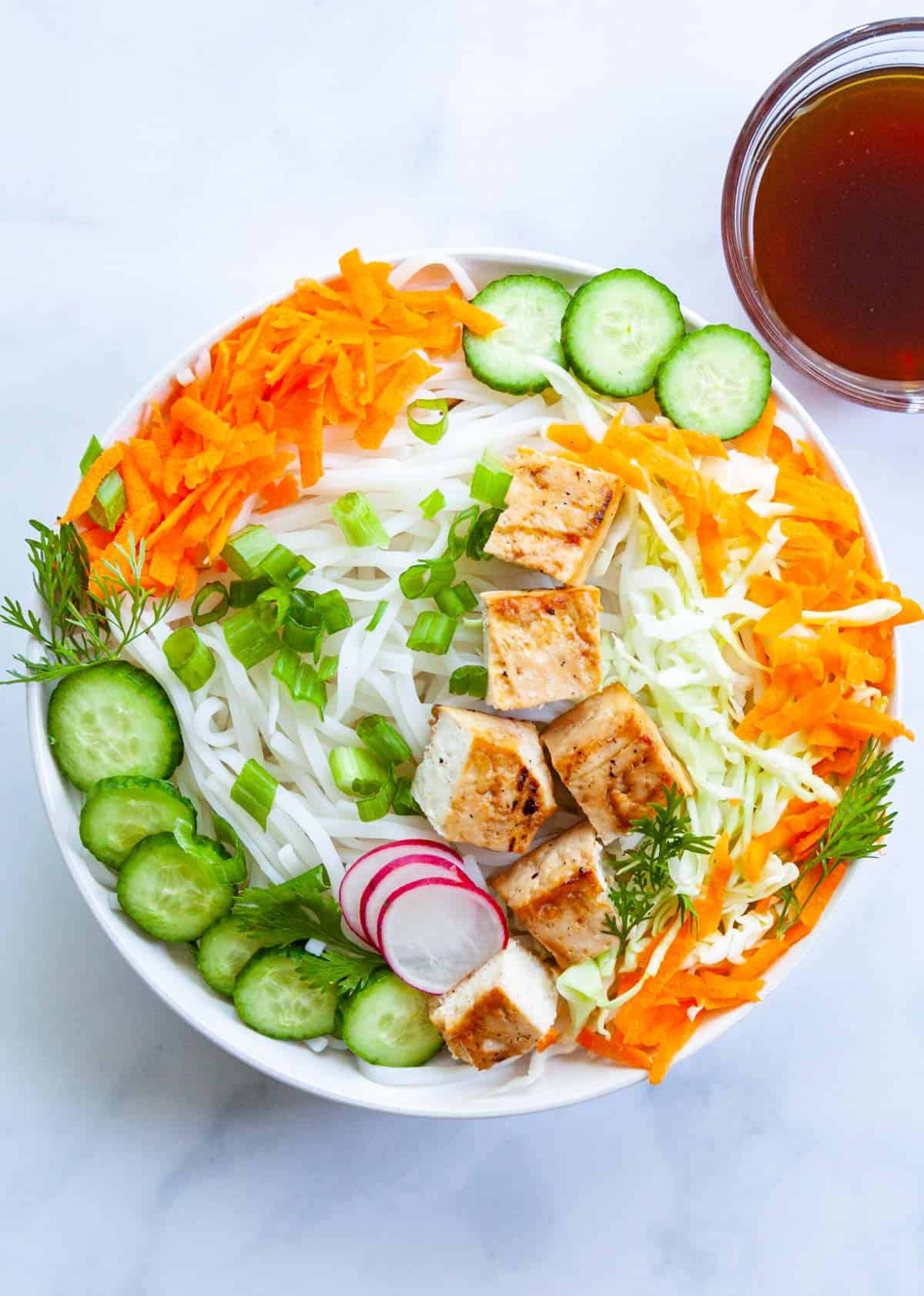 a bowl of rice noodles cucumber, cabbage, carrots and tofu next to a small bowl of soy sauce dressing