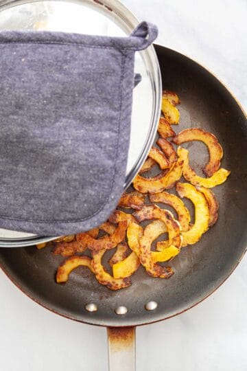 A lid being placed over a saute pan with slices of winter squash.